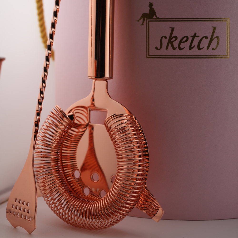 Load image into Gallery viewer, Rose gold cocktail strainer and spoon with 'sketch' branding on a pink background